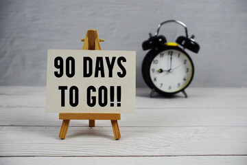 90 Days To Go! text with easel and alarm clock on wooden background
