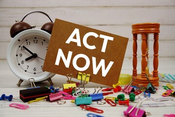 Act Now text on paper card with alarm clock and stationary on wooden background