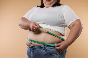 Overweight woman measuring her belly with tape on beige background, closeup