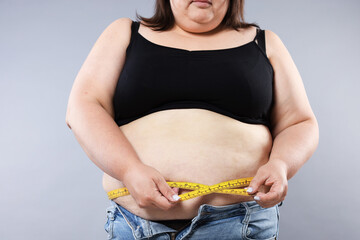 Overweight woman measuring her belly with tape on grey background, closeup