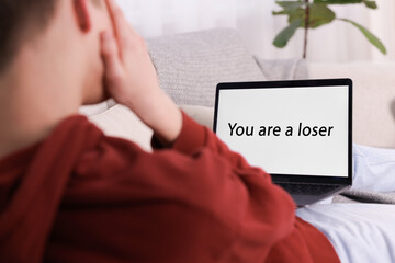 Cyber bullying. Upset teenage boy looking at laptop with text You Loser on sofa at home, closeup