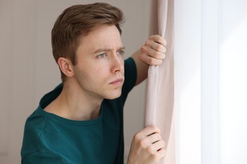 Paranoia. Worried man looking out window indoors