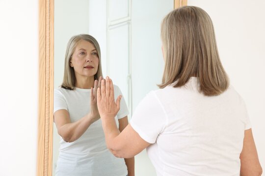 Beautiful senior woman looking at herself in mirror indoors - Powered by Adobe