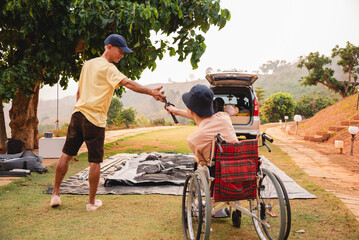 A young man with disability in wheelchair look at asian adult man setting up a camping tent, He...