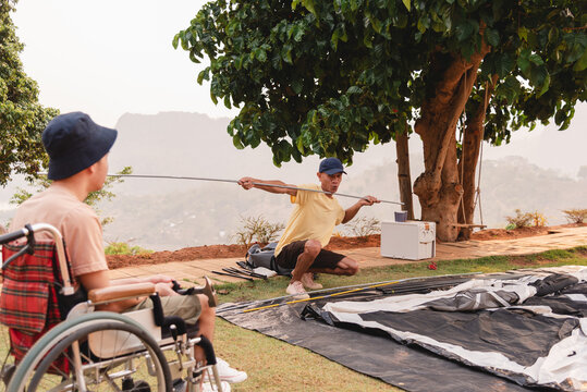 Asian adult man setting up a camping tent, he talks with a disability young man in a wheelchair with a smile on his face, A diversity family outdoor activity, Health mental, Father-son bond concept.