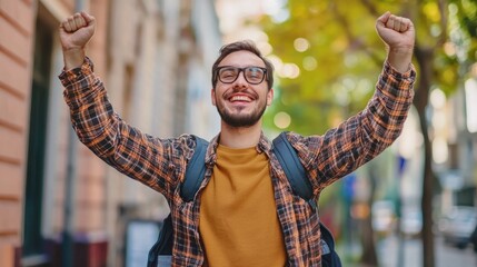 A man with glasses and a beard, wearing a plaid shirt and a backpack, standing outdoors with his arms raised in celebration.