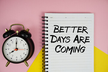 Better Days Are Coming text written on notepad with alarm clock on pink background