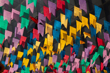 Bandeirolas festivas, coloridas, penduradas dançando ao vento com janelas de um predio desfocado...
