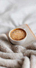 Creamy coffee in a white mug, resting on an open book, nestled in a soft beige blanket
