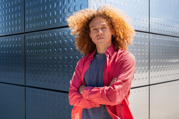 Young man with curly hair and pink shirt posing with arms crossed in front of metallic wall....