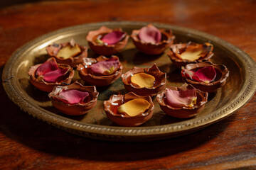 Multiple small clay diyas arranged in a circular pattern on a vintage brass tray, scattered rose petals and mango leaves between them, placed on a polished wooden table, rich festive texture