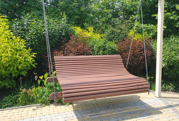 A wooden garden swing on metal chains in a sunny yard, surrounded by green bushes and ornamental...