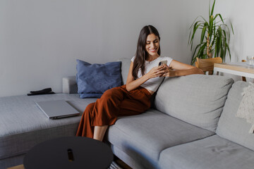 Smiling young woman using mobile phone while sitting on comfortable sofa in living room with laptop nearby