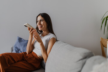 Happy young woman recording voice message on mobile phone while sitting on comfortable sofa in...