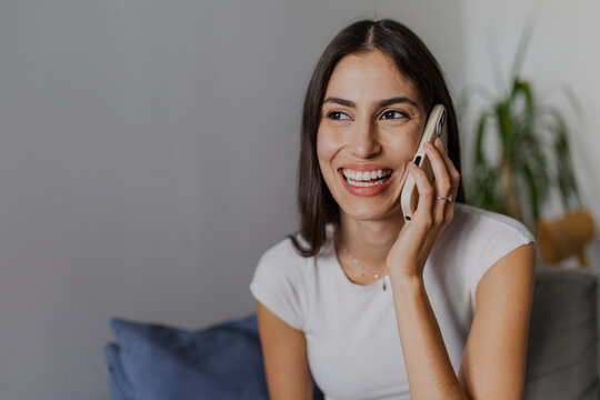 Happy young woman talking on mobile phone sitting on comfortable sofa at home, enjoying pleasant conversation