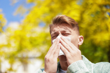 Young man with paper tissue suffering from seasonal pollen allergy near tree on spring day. Space...