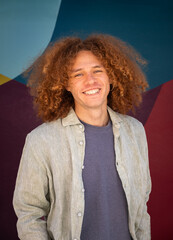Smiling young man with curly hair wearing linen shirt standing in front of colorful mural. Friendly attitude, urban fashion, modern youth, creative lifestyle and vibrant street background.