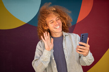 Curly-haired student waving during video call on smartphone, in front of vibrant urban mural. Youth lifestyle, street fashion and digital communication © LaMorenita