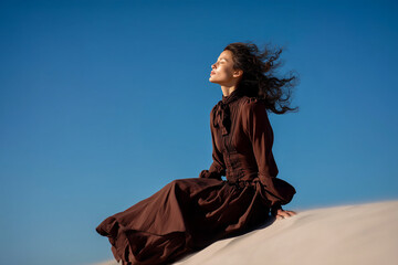 A woman in a brown dress is sitting on a sandy hillside, looking up at the sky
