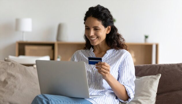 Happy Hispanic young woman consumer holding a credit card and a laptop while buying online at home. Female shopper customer shopping on an e-commerce website market place making a digital payment.