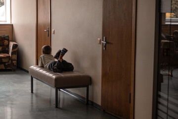 A young boy lies on a bench in a dim hallway, suggesting boredom, waiting, or a moment of quiet...