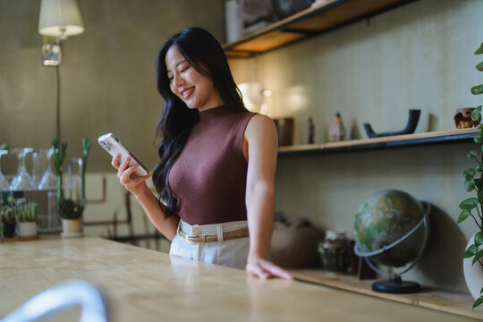 Asian businesswoman using mobile phone and smiling while leaning on wooden counter in cafe