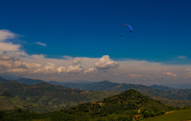 Parapente sobre colinas verdes y montañas con cielo azul profundo y nubes blancas en un día claro
