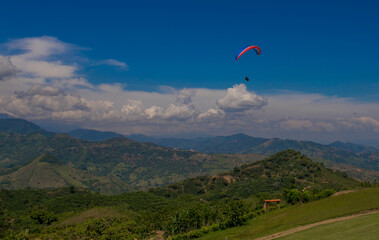 Parapentista volando sobre colinas verdes y montañas andinas en un día soleado con cielo azul y nubes blancas