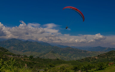 Parapentista en vuelo tándem sobre un paisaje montañoso verde bajo cielo azul con nubes blancas...