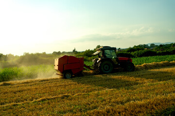 Agricultural scene with a red tractor and hay baler operating in a sunset field during harvest...