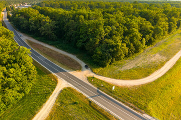 Drone image of a paved road intersecting with dirt paths near a lush forest. A place for a car to...