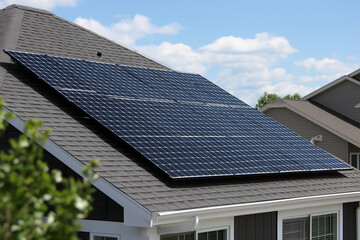 A solar panel sits tiled roof with white trim, catching sunlight from the upper left, while a neighboring gray-roofed house appears in front view.