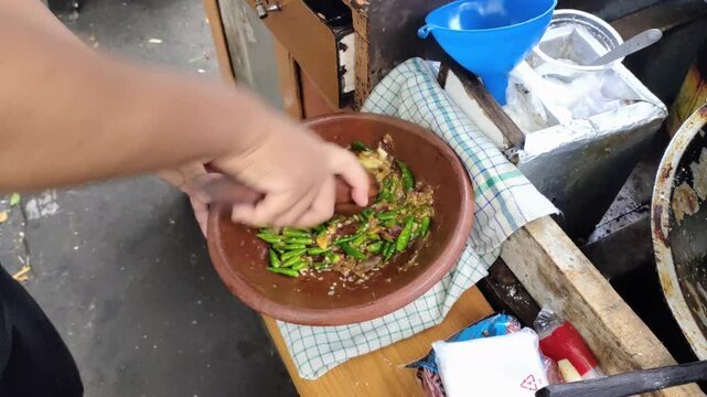 A tofu gejrot seller is pounding a mixture of green chilies, shallots, garlic and salt to make chili sauce for fried tofu gejrot. Tofu gejrot is a popular street food on the island of Java, Indonesia.