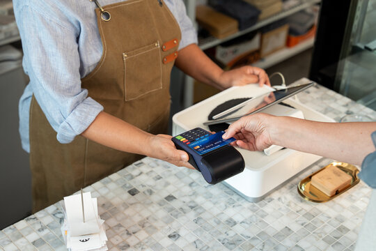 Customer inserting blue credit card into payment terminal held by barista in brown apron over marble counter at cafe during chip-based transaction using electronic point of sale machine