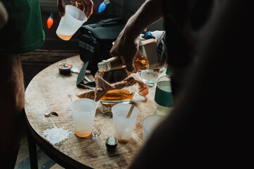 Hand pouring liquor into plastic cup on rustic wooden table with assorted bottles and orange juice...