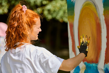 Young woman with curly red hair painting bright yellow sun on large canvas outdoors. Wearing white...