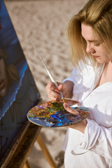 Young woman artist stands on sandy beach, engrossed in her painting. She holds colorful palette and...