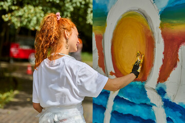 Young woman with curly red hair painting bright yellow sun on large canvas outdoors. Wearing white...
