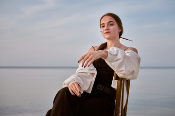 Young female artist in contemplative pose sits by calm ocean. Her serene expression and soft...