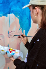 Young female artist with long hair painting portrait using vibrant blue hues. Focused expression, black sweater, baseball cap. Natural light illuminates canvas