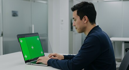 Focused young man works on laptop with green screen, modern office setting.