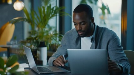 Focused african businessman multitasking with two laptops in a modern office space - Powered by Adobe