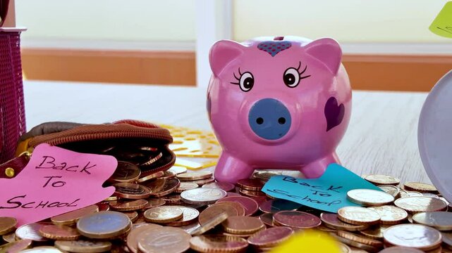 Pale hand carefully inserting euro coin into pink piggy bank, surrounded by scattered coins and educational labels, representing financial planning for school expenses