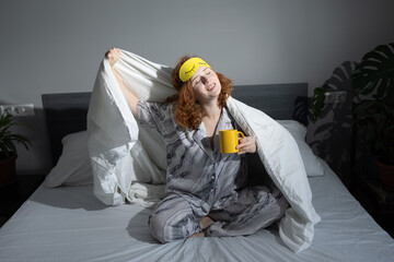 A young woman in gray pajamas sits on her bed, stretching with a blanket over her shoulders while...
