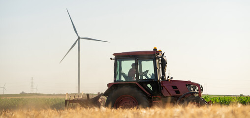 Agricultural tractor is working on the field with wind turbines