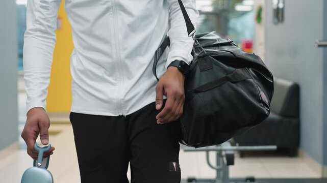 close up of athlete carrying black duffel bag and wireless headphones walking through bright modern gym past weight bench and equipment reflecting in glass as natural light filters through windows