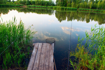 Scenic view of a calm lake with fishing rods and a net on a wooden pier. Peaceful countryside...