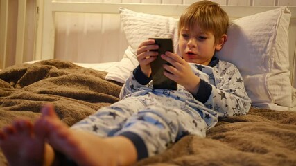 Cute kid boy in pajamas lying in bed watching smartphone, smiling. Children's screen time. Child plays games using phone at the evening or night. Social media addiction. Childhood, modern technology - Powered by Adobe