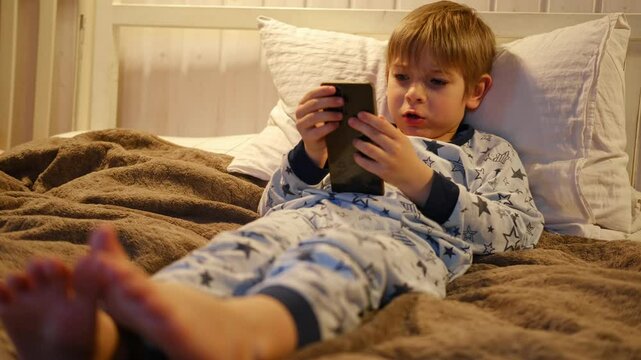 Cute kid boy in pajamas lying in bed watching smartphone, smiling. Children's screen time. Child plays games using phone at the evening or night. Social media addiction. Childhood, modern technology