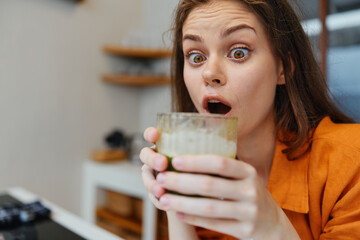 Surprised young woman holds a glass of healthy green smoothie, expressing delight and curiosity in...